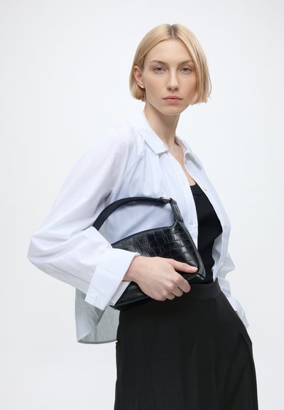 Jeune femme aux cheveux blonds courts, portant une chemise blanche et un pantalon noir, tient un sac à main noir texturé sur un fond blanc uni.