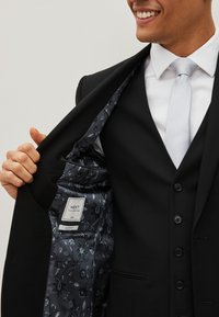Black tailored suit with a silver tie. Interior features a dark floral pattern and label. Slim fit design, with six buttons visible.