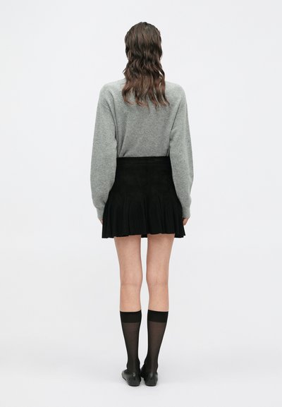 Grauer Pullover, schwarzer Rock mit plissierter Textur, transparente schwarze Socken und schwarze flache Schuhe. Von hinten vor einem hellen Hintergrund aufgenommen.