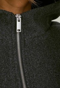 Giacca grigia con cappuccio realizzata in un tessuto testurizzato e felpato. Presenta una cerniera metallica frontale e un design con collo alto.