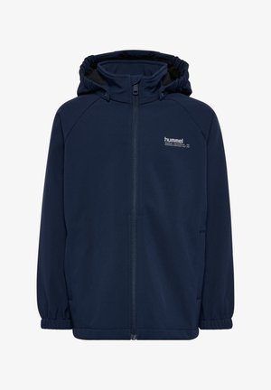 Junges rothaariges Mädchen, das eine marineblaue Jacke, ein graues Hemd, dunkle Jeans und blaue Sneakers trägt, steht vor einem schlichten hellen Hintergrund.