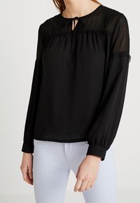 Schwarze Bluse mit einem transparenten gepunkteten Einsatz und rüschigen Akzenten. Mit einem gebundenen Ausschnitt und elastischen Bündchen, kombiniert mit hellen Hosen.