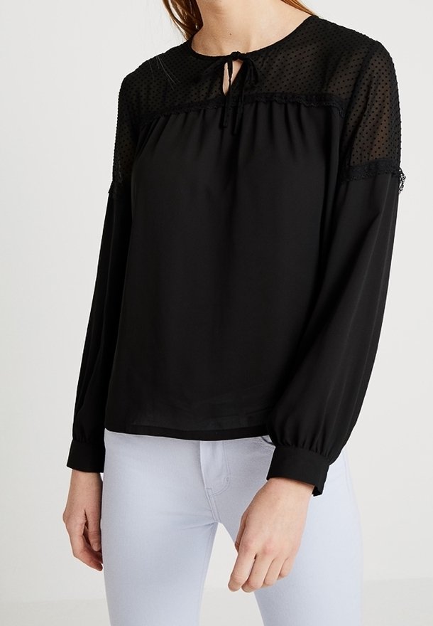 Schwarze Bluse mit einem transparenten gepunkteten Einsatz und rüschigen Akzenten. Mit einem gebundenen Ausschnitt und elastischen Bündchen, kombiniert mit hellen Hosen.