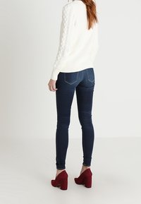Suéter blanco de punto con patrón texturizado, jeans ajustados azul oscuro y zapatos de tacón rojos. Vista desde la parte de atrás.