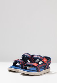 Blå och röda sandaler med justerbara remmar, tillverkade av textil och gummi. Har en lätt sula och en öppen tå-design för bättre andningsförmåga.