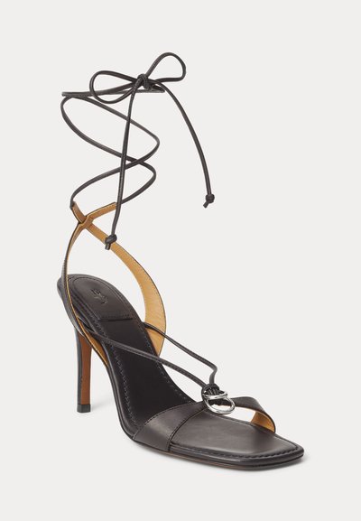 Μαύρο δερμάτινο σανδάλι με ψηλό τακούνι και τετράγωνη μύτη, λεπτά λουράκια που διασταυρώνονται πάνω στο πόδι, ασημένια πόρπη και κορδέλες που δένουν γύρω από τον αστράγαλο σε φιόγκο.
