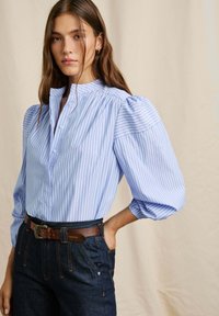  Lichtblauwe gestreepte blouse met pofmouwen, knoopsluiting bij de kraag en textuurstof, gecombineerd met een donkere spijkerbroek en een bruine riem.