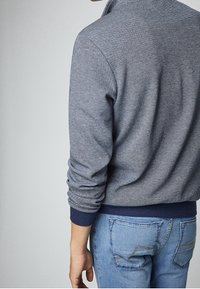 Suéter texturizado de color azul marino con cuello alto y puños acanalados, combinado con jeans de color azul claro que tienen bolsillos traseros y detalles cosidos.
