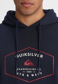 Námořnická modrá mikina s kapucí s hexagonálním logem v bílé a červené barvě, s tkanicemi na stažení a klokankou. Měkká textura, ležérní design.