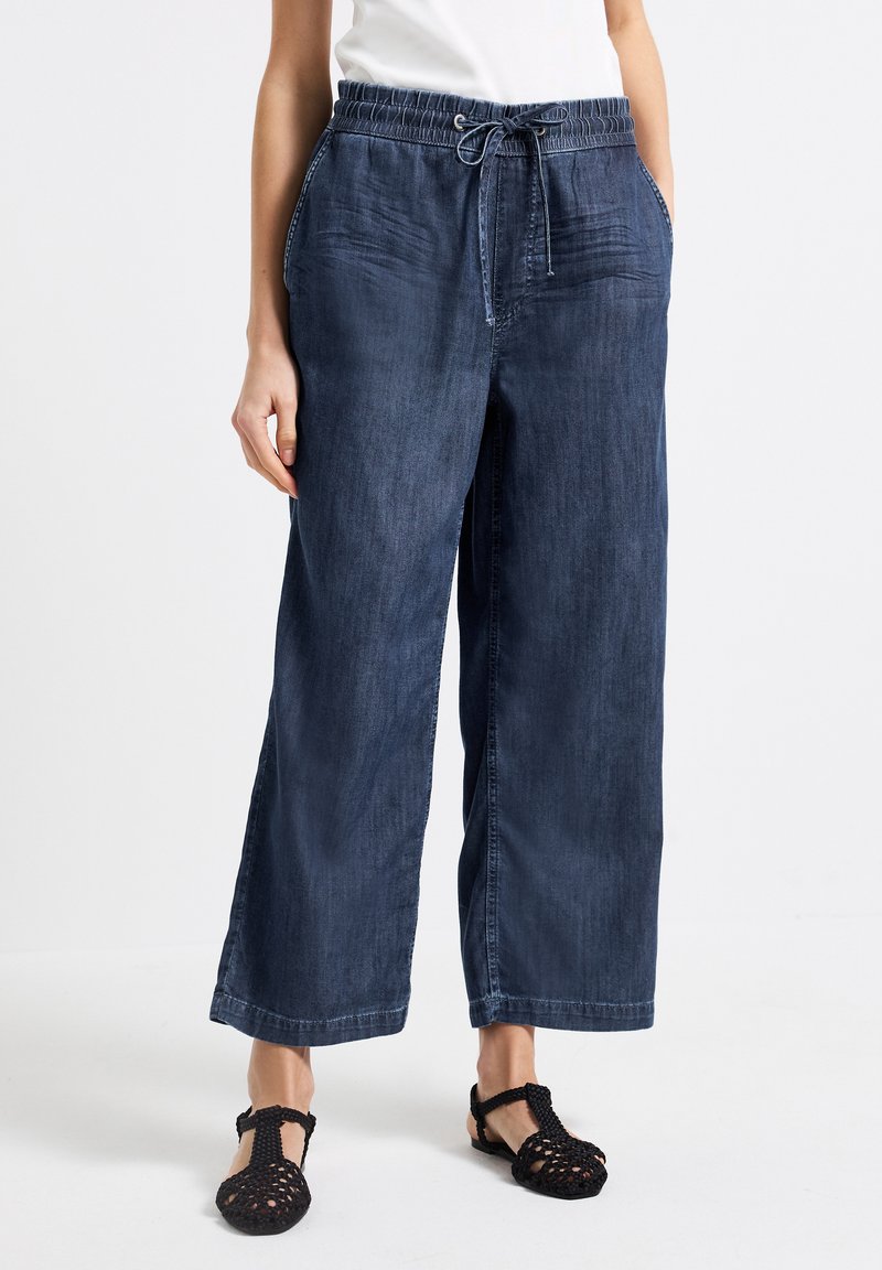 Široké modré džíny s elastickým pasem a šňůrkou na stažení. Obsahují boční kapsy a hladkou texturu. Spárovány s černými sandály.