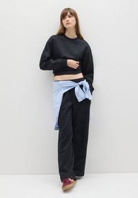 Schwarzes, gekürztes Sweatshirt mit Sprenkeln, kombiniert mit schwarzen Loungehosen und einem hellblau gestreiften Shirt, das in der Taille gebunden ist. Burgunderfarbene Sneaker.