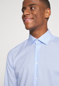 Camicia azzurro chiaro a maniche lunghe con colletto a punta e patta frontale. Tessuto liscio con un design aderente e bottoni trasparenti.