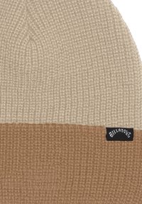 Gorro de punto en dos colores: parte superior color beige claro y parte inferior marrón. Presenta un patrón texturizado y una etiqueta negra con texto blanco.
