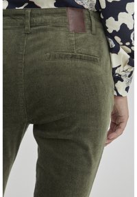 Pantaloni in velluto a coste verde con rigature verticali, dotati di una toppa in pelle marrone sulla vita posteriore e una singola tasca sul retro.