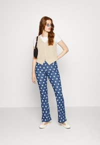 Colete de malha bege sobre uma t-shirt branca, emparelhado com jeans flare azuis com padrões florais brancos e ténis brancos. Mala preta.