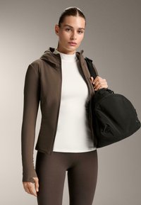 Hnedá ľahká bundy na zips s kapucňou, kombinovaná s bielym topom s vysokým golierom a hnedými legínami. V ruke drží čiernu kabelku zaobleného tvaru.