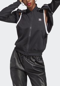 Veste noire zippée avec des accents blancs et un design à épaules découpées. Fabriquée en tissu lisse, elle dispose de poches latérales et de poignets côtelés.