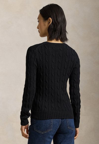 Nainen, jolla on lyhyet tummat hiukset, pukeutuneena mustaan palmikkoneulepaitaan ja sinisiin farkkuihin, katsomassa pois päin neutraalia taustaa vasten.