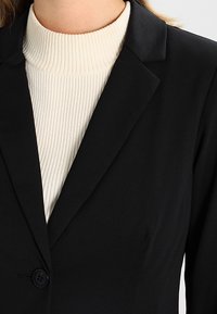 Czarna marynarka z klapami, zapinana na jeden guzik, z gładkiego materiału, nałożona na białą sweter z prążkowanym golfem.