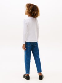 Hvit langermet skjorte, blå denim jeans med lett falming, svarte slip-on sko; krøllete hår rammet inn ved skuldrene.