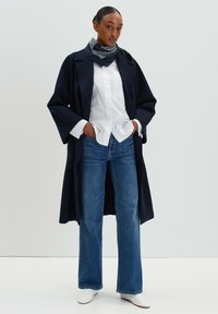 Marineblauwe jas met brede mouwen over een witte blouse, gecombineerd met blauwe flared jeans en een gestreepte sjaal, met witte schoenen.