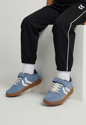 Osoba siedząca ubrana w czarne spodnie dresowe i białe skarpetki, prezentująca niebieskie trampki z białymi paskami w kształcie litery V i brązowymi podeszwami.