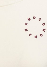 Crème-kleurige sweatshirtstof met een textuur. Heeft een cirkelvormig bordeauxrood logo met letters die rondom de cirkel zijn uitgelijnd.