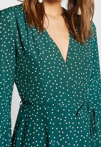 Vestido verde con un escote en V profundo, mangas largas y lunares blancos. Tiene un lazo en la cintura y una falda fluida.