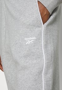 Main dans la poche d'un pantalon de survêtement Reebok gris clair avec un liseré blanc le long de la couture latérale et un logo sur la cuisse.