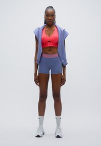 Αθλητικό σουτιέν σε ροζ με το λογότυπο «Nike Pro», συνδυασμένο με λιλά σορτς και ένα ελαφρύ μωβ αμάνικο φούτερ με κουκούλα. Λευκές κάλτσες και παπούτσια.