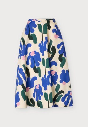 Femme portant un pull duveteux rose clair à col en V, une jupe midi crème avec des motifs floraux bleus et verts, et des talons pointus noirs.