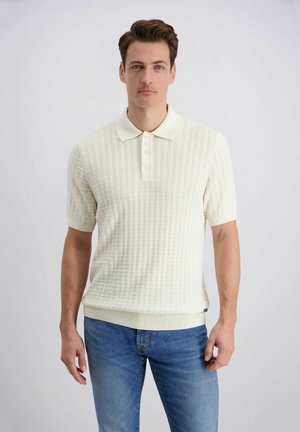 Homme aux cheveux bruns courts portant un polo crème à manches courtes texturé et un jean bleu, debout devant un fond clair uni.