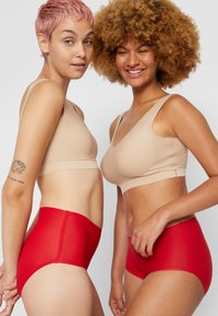 Dvě modelky mají na sobě béžový hladký bralette a červené kalhotky s vysokým pasem. Bralette má výstřih do V a široká ramínka; kalhotky jsou hladké a přiléhavé.
