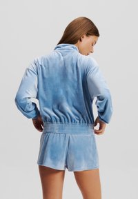 Ljust blå velourlounge-set som innehåller en zip-up jacka med ståkrage och shorts med elastisk midja, med en mjuk textur och avslappnad passform.