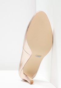 Blekguld klackskor med en slät, glänsande yta, spetstå, och en smal klack; har en texturerad gummisula och synlig varumärkeslogotyp.