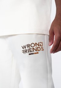 Witte katoenen t-shirt met een ronde halslijn. Daarnaast witte broek van textuurstof met het geborduurde logo "WRONG FRIENDS" in bruin.
