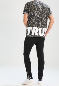 Kameramönstrad kortärmad skjorta med vit "TRUE" text, tillsammans med svarta tapered byxor med dragkedjor vid anklarna och vita sneakers.