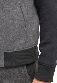 Grijze en zwarte fleece sweatshirt met een zijzak en ribgebreide manchetten, met een klein merkmetalen accent nabij de zoom.