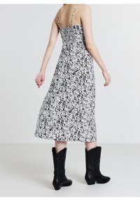 Robe maxi blanche à pois noirs avec bretelles spaghetti, dos froncé, tissu fluide, portée avec des bottes de cowboy noires.