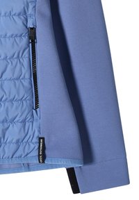 Blå jakkeærme og quiltet sidepanel med sort lynlås og "Cecil Outerwear"-label synlig på stoffkanten.