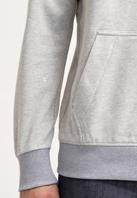Gros plan d'une personne portant un sweat-shirt gris clair avec des poignets côtelés et une poche avant sur le côté.