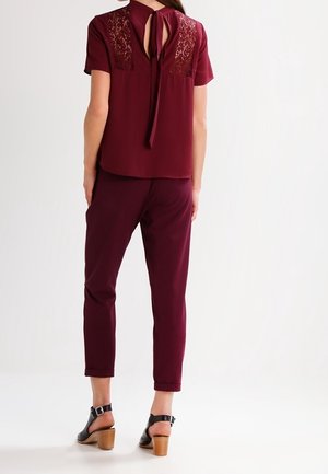 Nainen, jolla on päällään viininpunainen toppi, jossa on pitsiset takapaneelit ja solmittava yksityiskohta, samanväriset lyhyet housut sekä mustat korolliset sandaalit nilkkaremmeillä.
