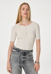 Lichtbeige geribde top met korte mouwen en een ronde halslijn, voorzien van zes decoratieve knopen aan de voorkant. Gemaakt in combinatie met grijze jeans.