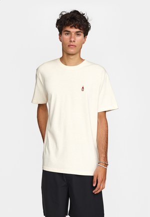 Jeune homme aux cheveux bouclés portant un t-shirt uni écru avec un petit logo rouge en forme de bouteille sur la poitrine et un short noir.