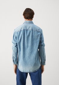 Light blue denim shirt with long sleeves, a classic collar, and a curved hem. Features visible seams and a relaxed fit.