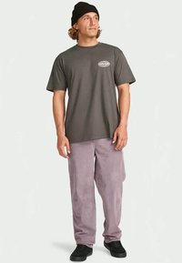 Mann, der eine schwarze Mütze, ein graues Graphic-T-Shirt, eine helllilafarbene Cord-Hose und schwarze Turnschuhe trägt, steht und nach links schaut.