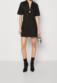 Schwarzes kurzärmliges Kleid mit tiefem V-Ausschnitt und einem runden Ausschnitt in der Taille. Kombiniert mit schwarzen Sock-Boots und einer kleinen silbernen Handtasche.