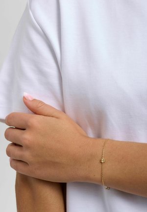 Personne portant un bracelet fin en or avec un petit pendentif en forme de fleur au poignet, habillée d'une chemise blanche à manches courtes unie, les bras croisés.