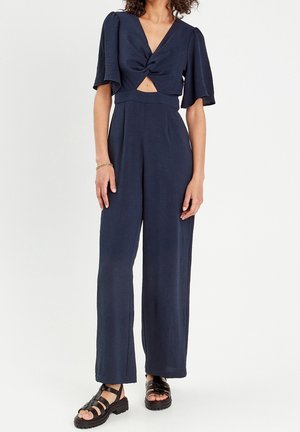 Femme portant une combinaison bleu marine à jambes larges, nouée à l'avant, avec une découpe à la taille, et des sandales noires à brides avec semelles épaisses.