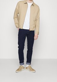 Chaqueta ligera beige, camiseta blanca, vaqueros azul marino y zapatillas beige con detalles blancos. Atuendo casual con un ajuste relajado.